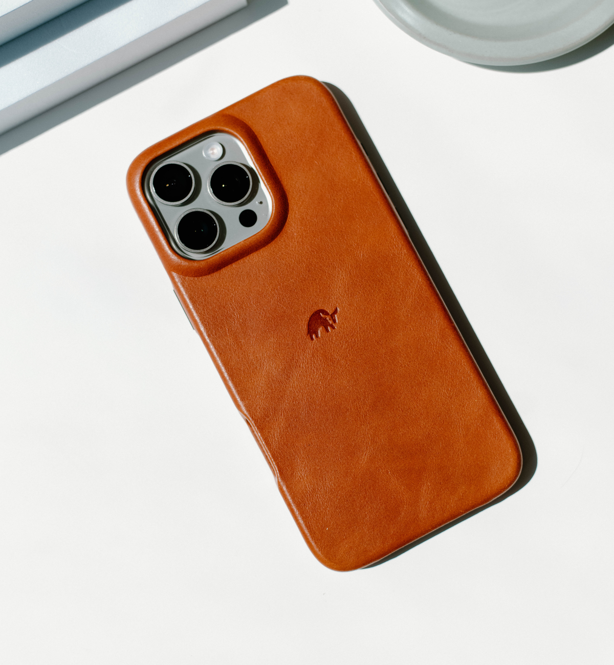 Brown Sienna leather iPhone case with embossed mammoth logo and raised camera cutout, angled on white surface.