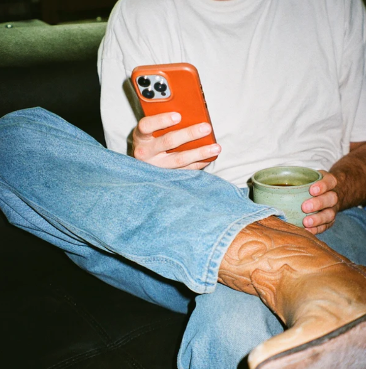 Person sitting cross-legged in jeans and brown cowboy boots, holding an orange smartphone and a green mug.