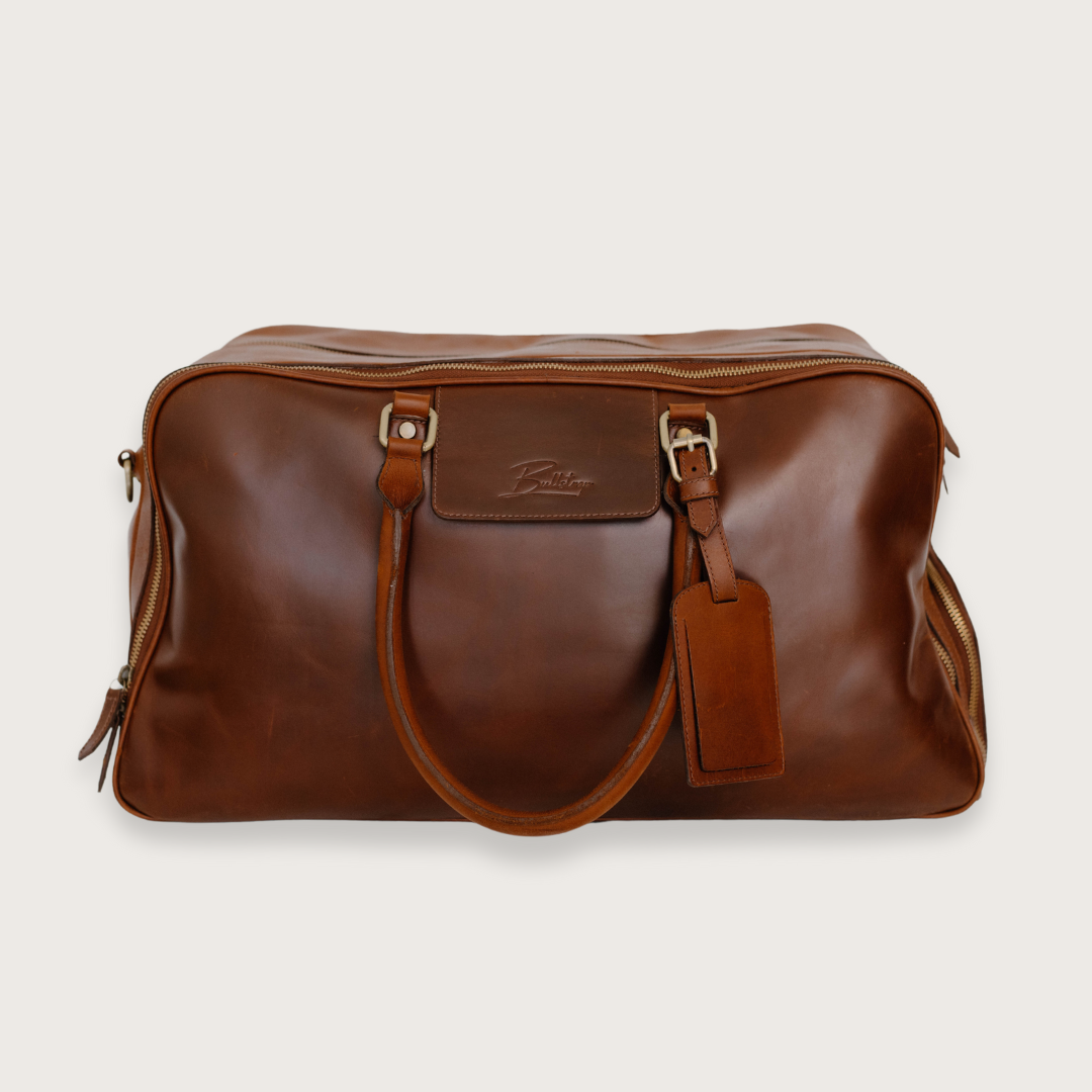 Brown leather travel bag with a luggage tag, labeled Bullstrap Reserve - The Odyssey Travel Bag