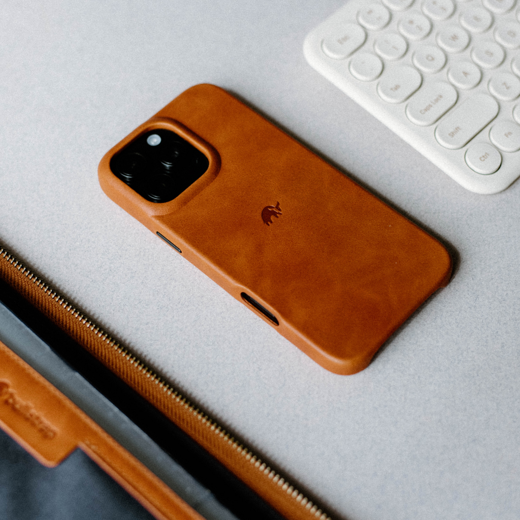 Sienna brown leather iPhone case with embossed elephant logo next to a white keyboard and zippered leather sleeve