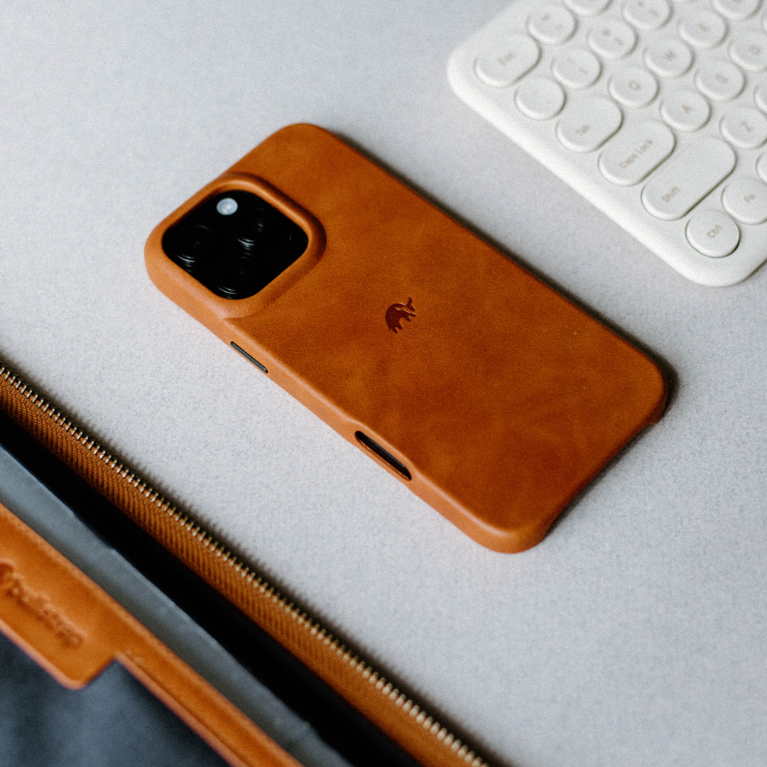 Sienna brown leather iPhone case with embossed elephant logo next to a white keyboard and zippered leather sleeve