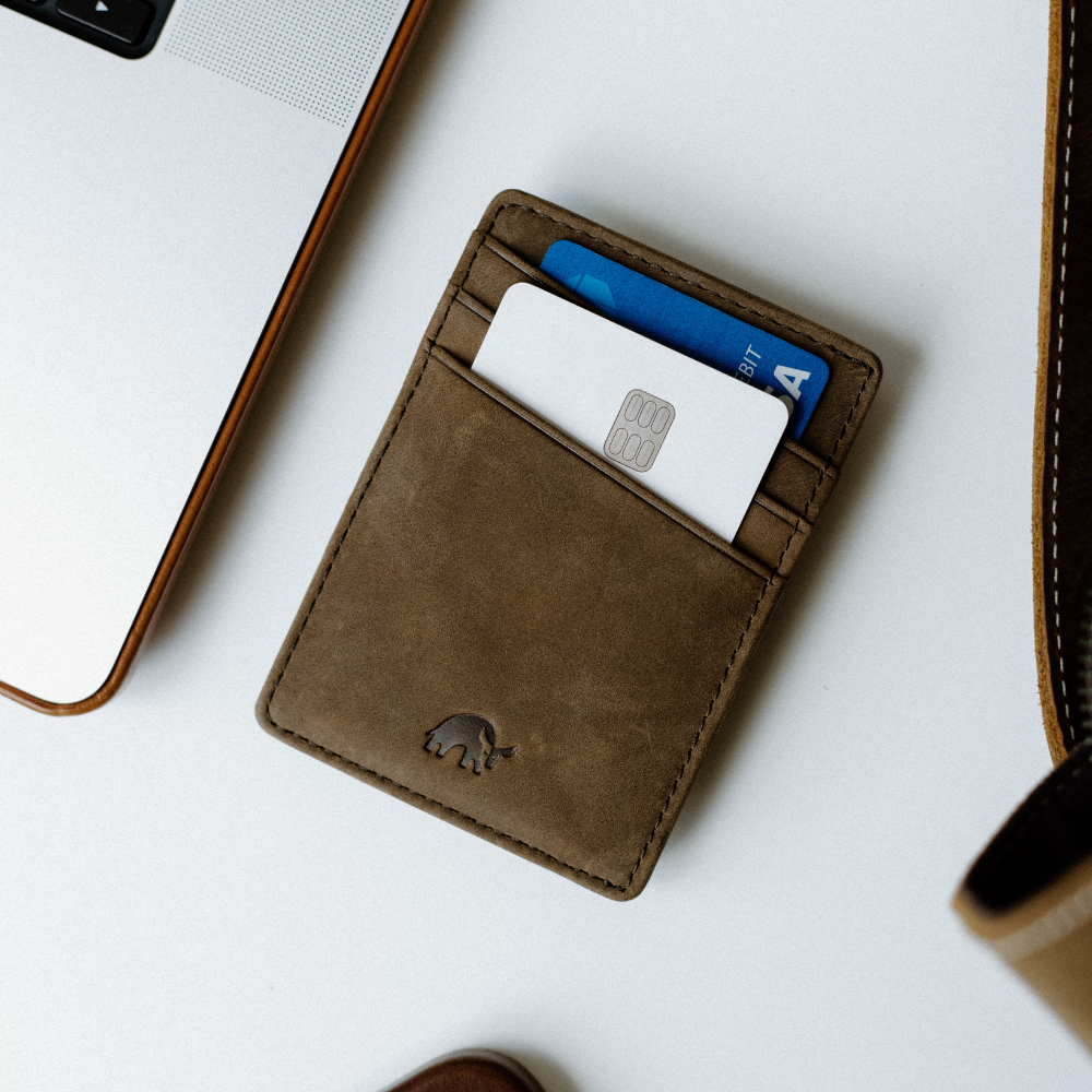 Brown leather card wallet on white desk with a white chip card and blue debit card showing DEBIT and VISA, laptop corner