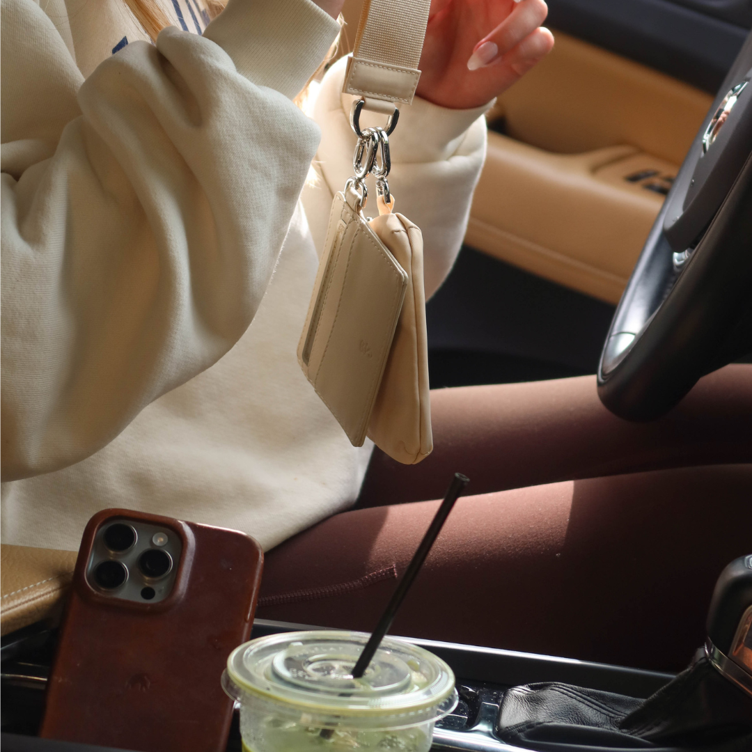 Hands holding beige cardholder and pouch on a strap in a car; phone on console and iced drink in cupholder.