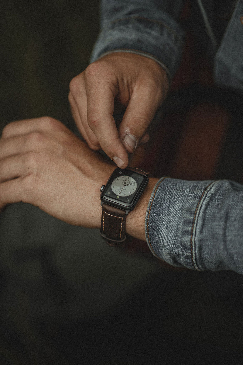 Hands adjusting a smartwatch with brown leather strap on a wrist, denim shirt cuff visible