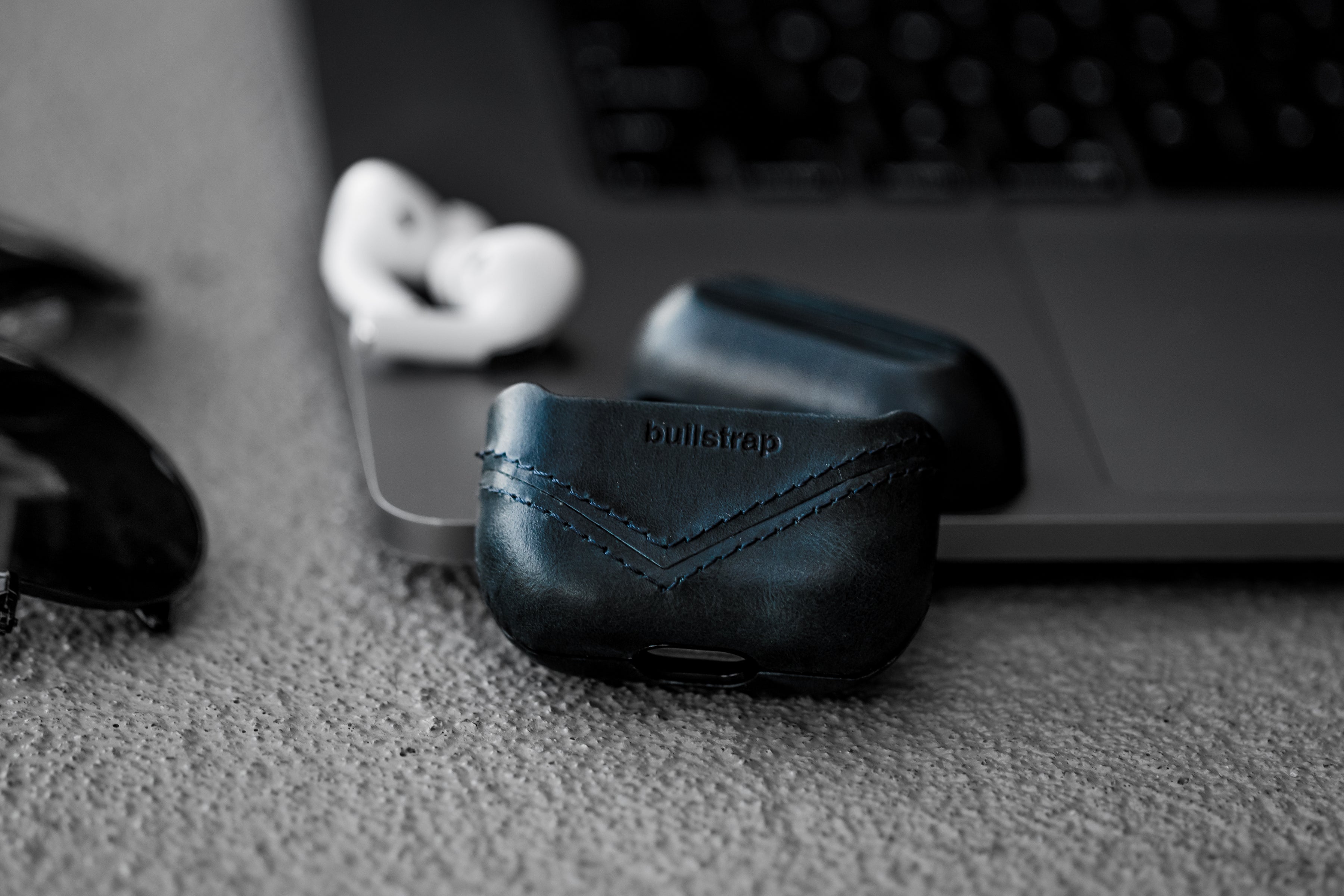 Black leather AirPods case embossed with 'bullstrap', white AirPods and laptop keyboard blurred in the background.