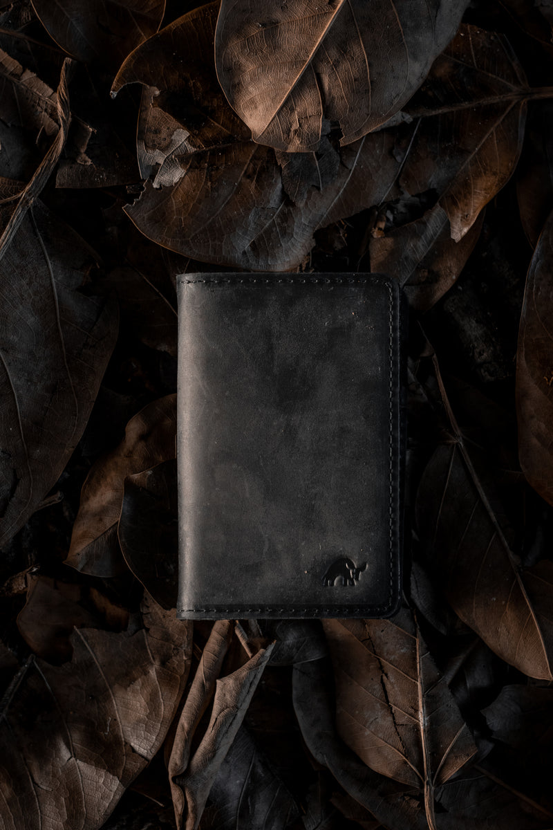 Black leather bifold wallet with small embossed elephant logo resting on dark, dried leaves.