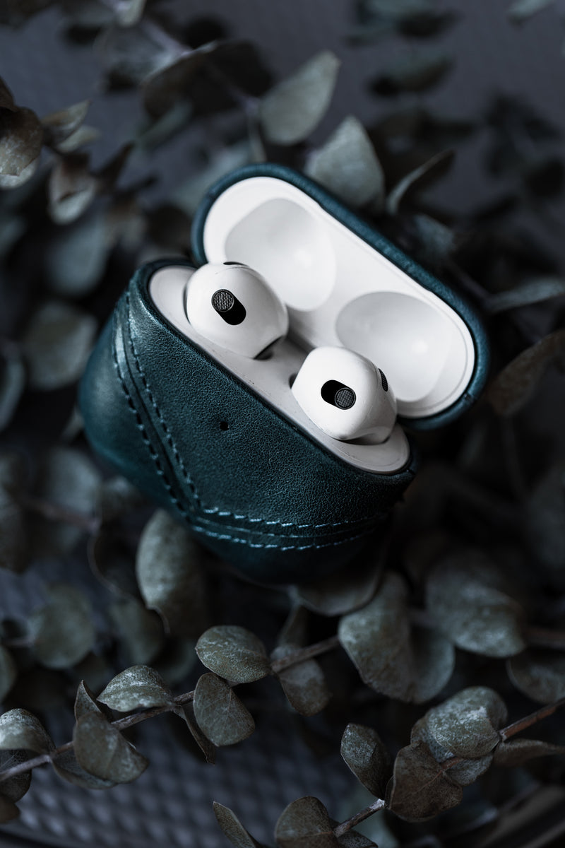 Leather AirPods case in teal color with AirPods inside, surrounded by leaves.