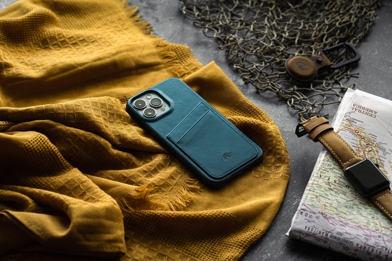 Teal iPhone with leather wallet case resting on mustard textured scarf beside a map labeled CENTRAL AMERICA and a leather-strapped watch.