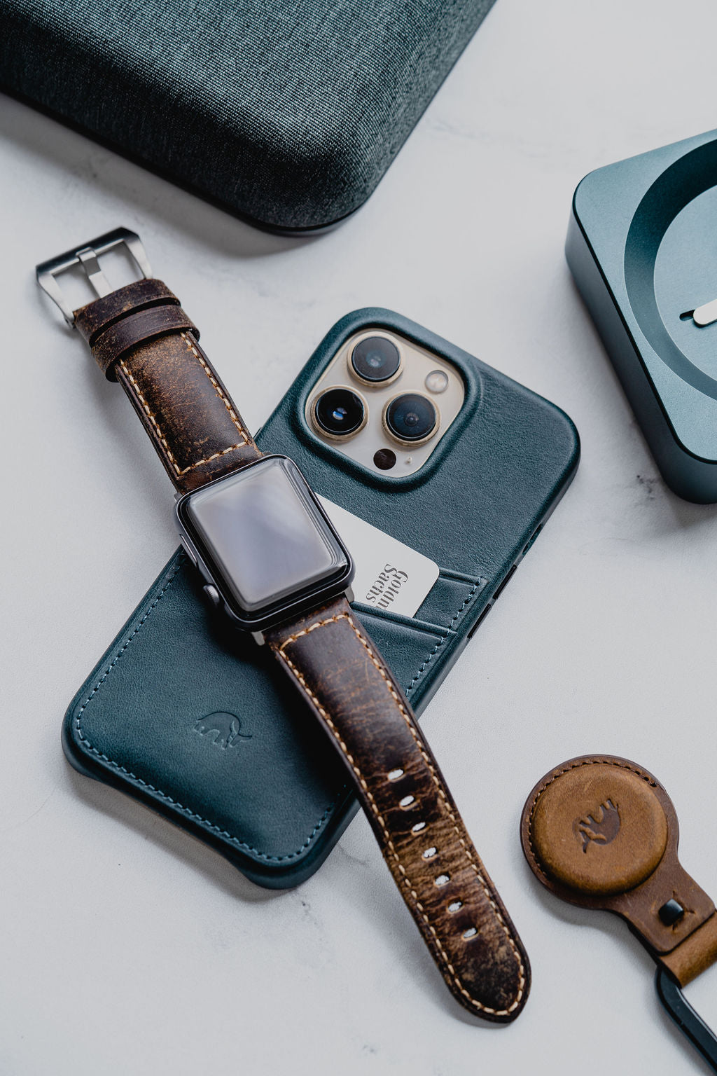 Apple Watch with brown leather band resting on a teal leather iPhone case with a pocketed card and leather AirTag holder.