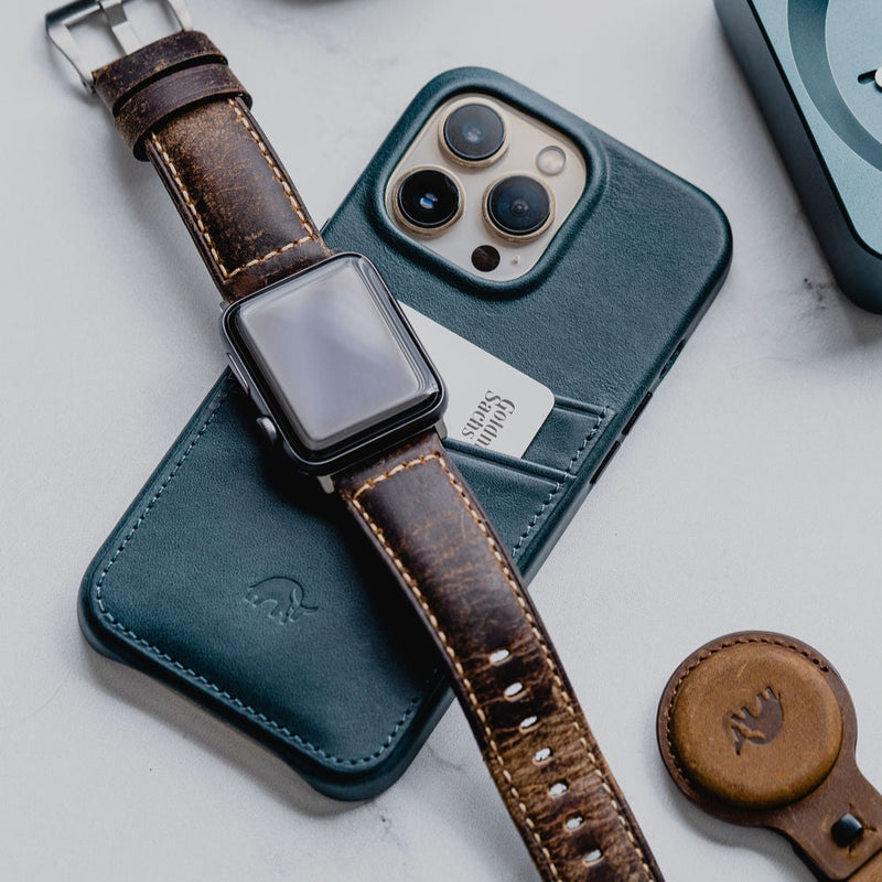 Teal iPhone case with card in slot; Apple Watch with brown strap across it; brown AirTag holder on marble.