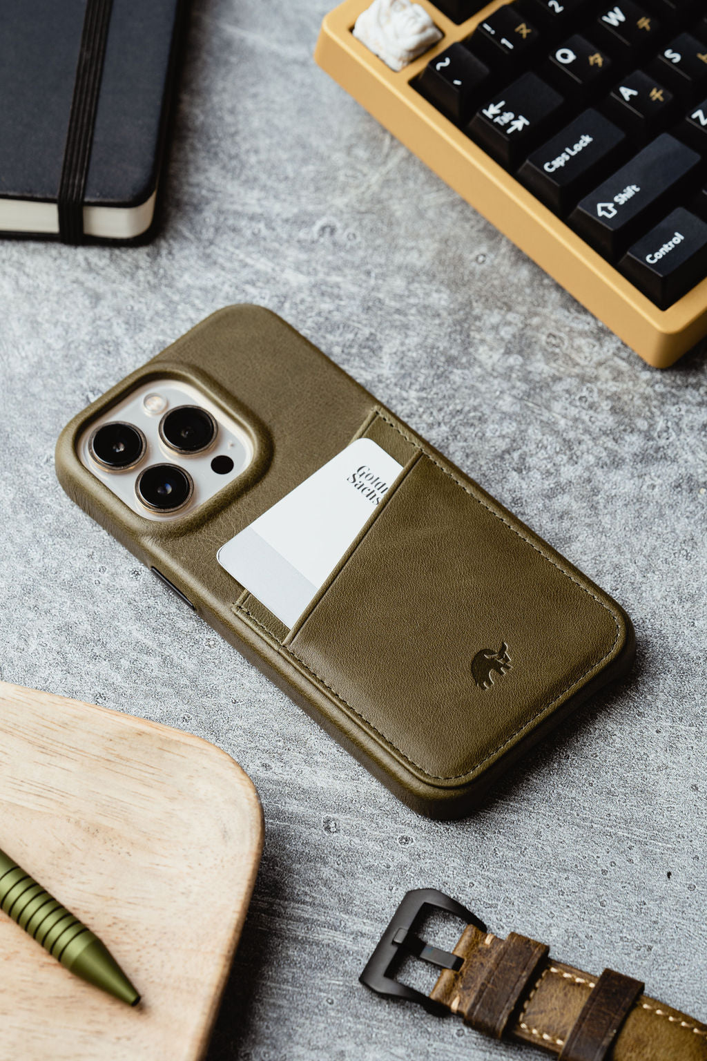 Olive green leather iPhone case with a card pocket holding a white card on a concrete desk beside a keyboard and watch strap.