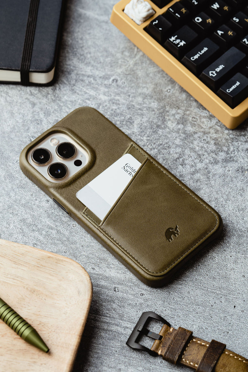 Olive green leather iPhone case with a card pocket holding a white card on a concrete desk beside a keyboard and watch strap.