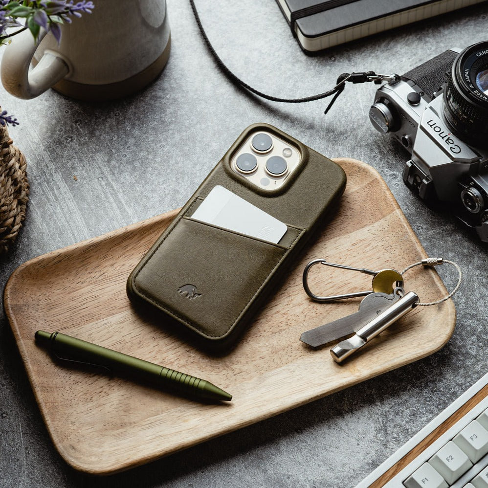 A green phone case with a card slot, keys, and a pen on a wooden tray.