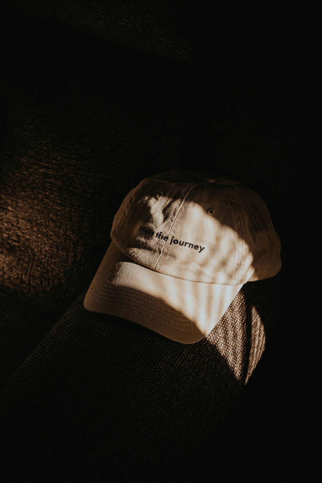 Beige baseball cap on dark couch with striped sunlight; embroidered words: enjoy the journey.
