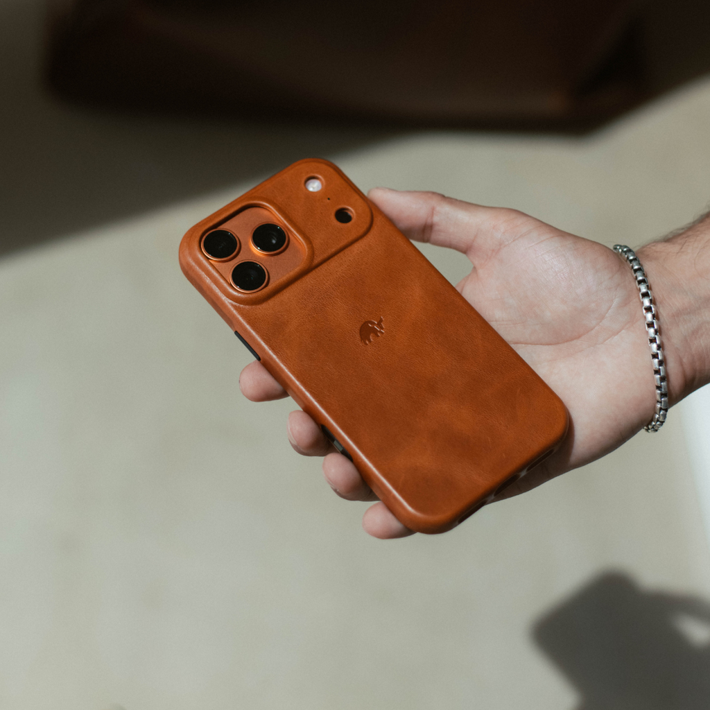 Brown leather iPhone case with triple camera cutout and small embossed logo, held in a hand wearing a chain bracelet