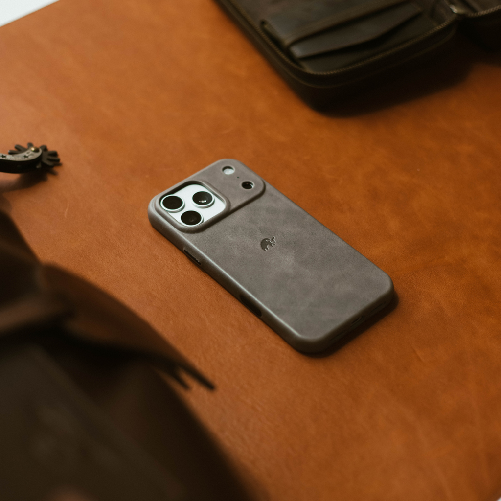 Slate leather iPhone case with triple-camera cutout and small embossed logo lying on a tan leather surface