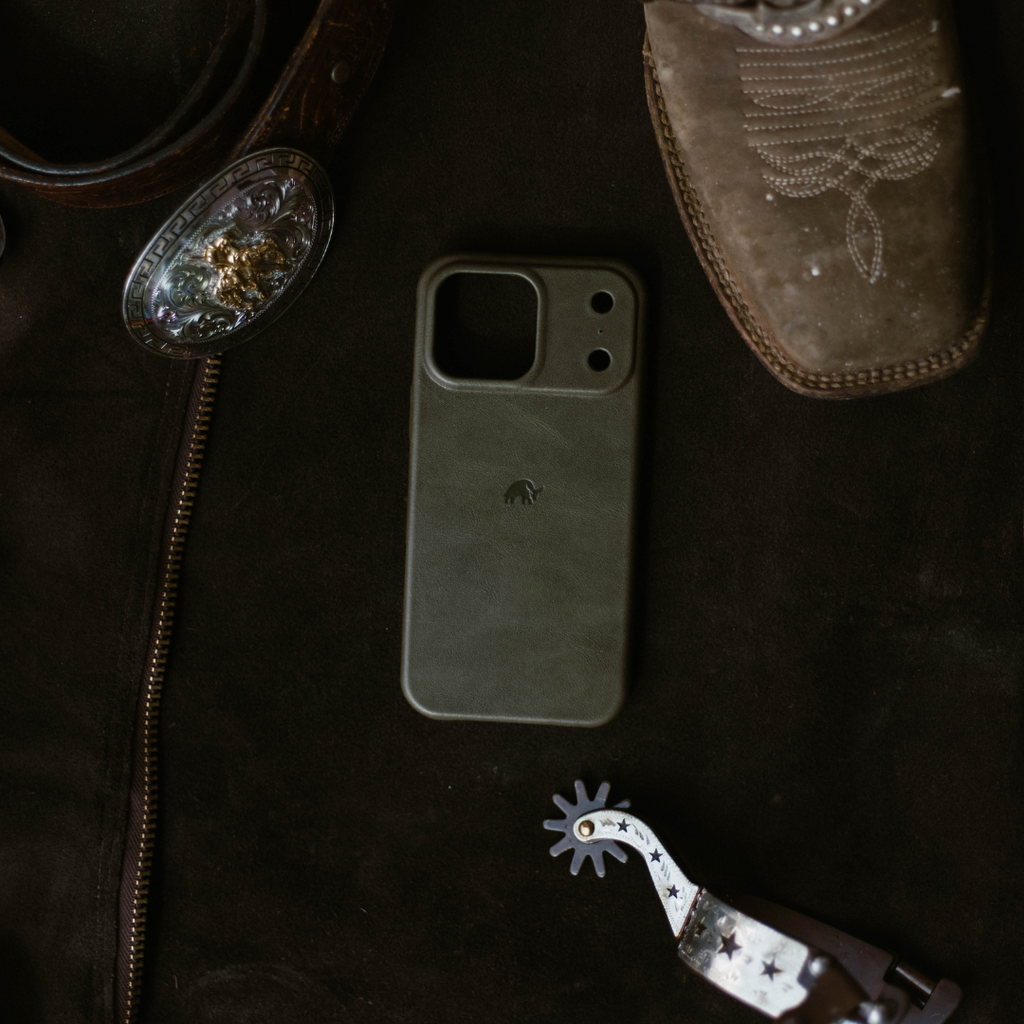 Green leather phone case with elephant logo on dark cloth beside a cowboy boot, spur, and decorative belt buckle.