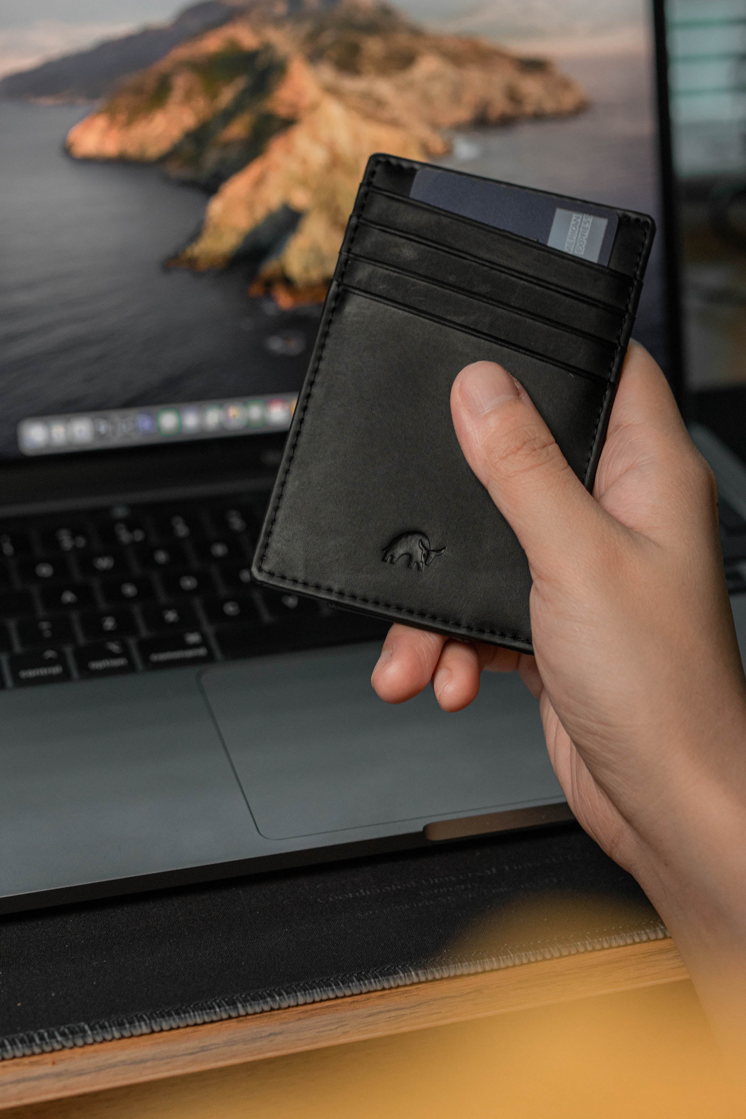 Black leather slim card wallet held above a laptop keyboard, thumb on the front card slot and a small embossed rhino logo.