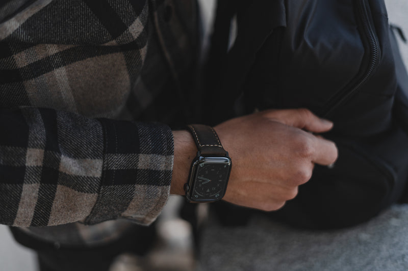 Wrist wearing a black smartwatch with leather band, plaid jacket sleeve; hand gripping a black backpack strap.