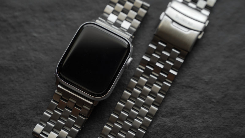 Silver smartwatch with black screen and two stainless-steel link bracelets laid on dark textured slate