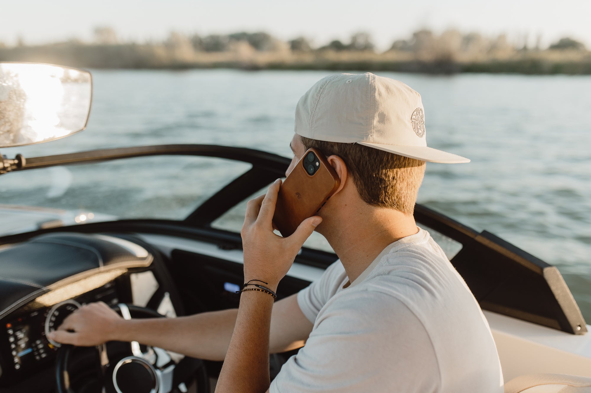 Young man wearing a beige cap steering a boat while holding a smartphone to his ear, lake and shoreline in background.