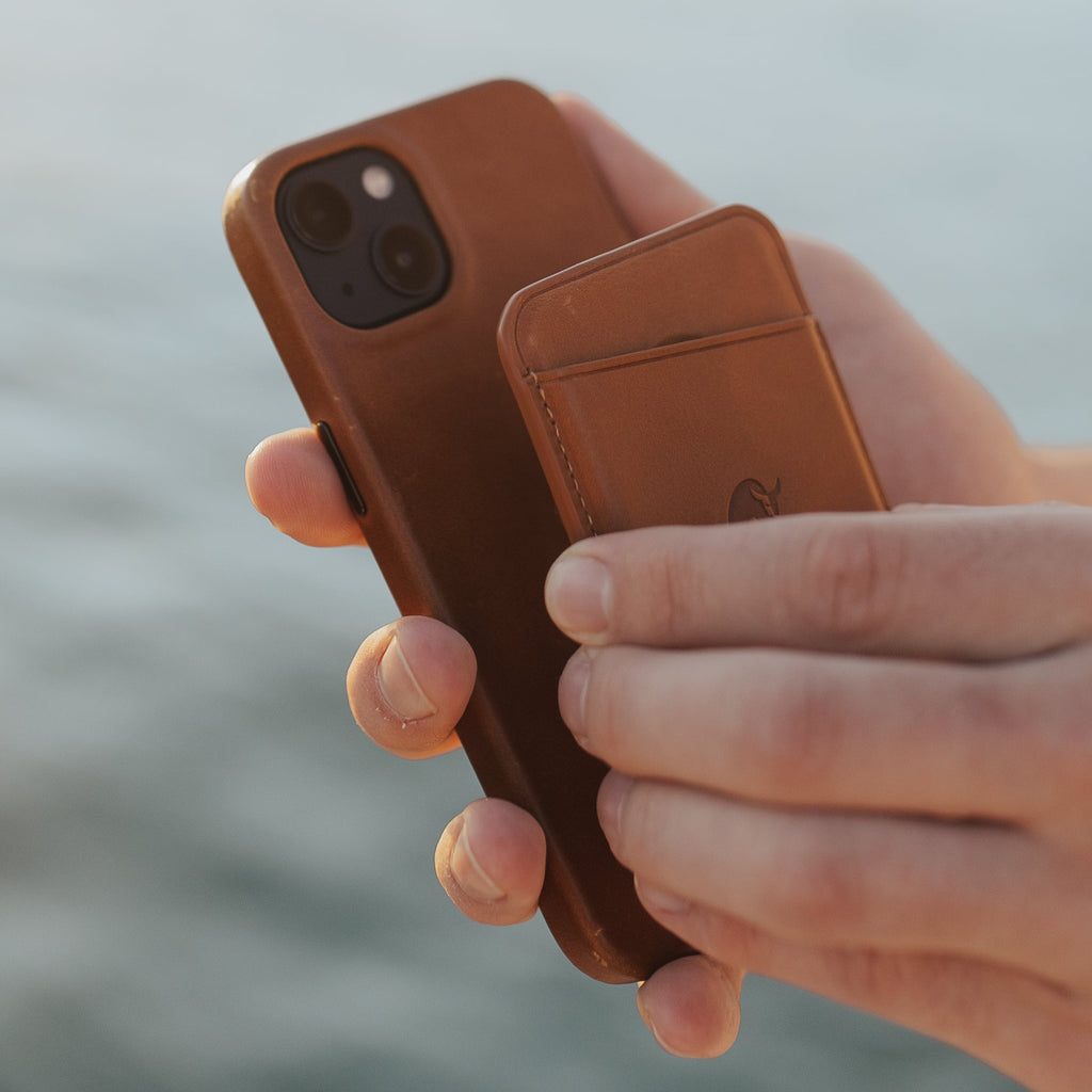 A person holding a brown leather magnetic wallet and iPhone by the water.