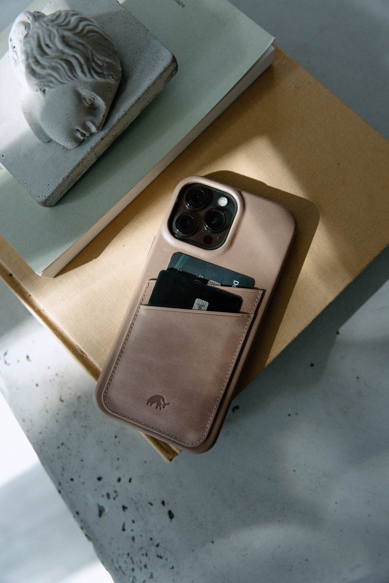 Bullstrap iPhone case in beige with cards in the back pocket, placed on a book and a surface.