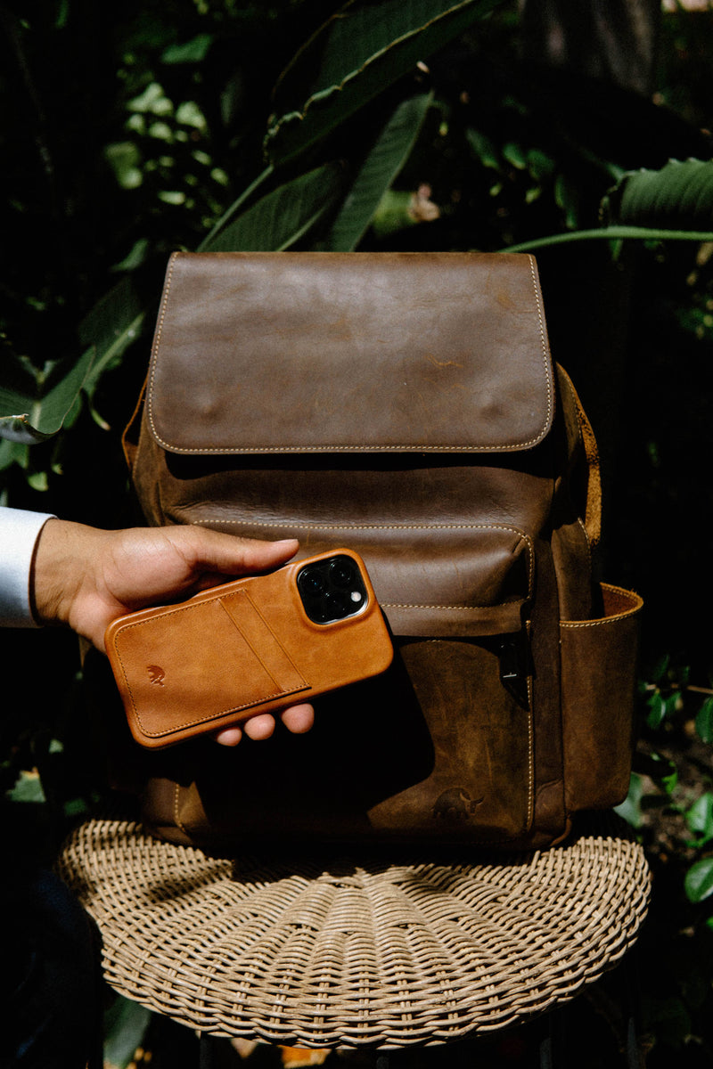 Hand holding tan leather iPhone case in front of brown leather backpack on wicker stool, leafy background.