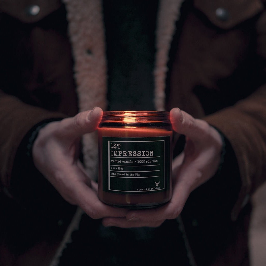 A person holding a candle labeled '1ST IMPRESSION', crafted with 100% soy wax, hand poured in the USA.