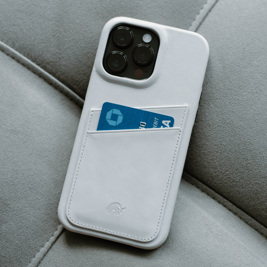 White leather iPhone case with stitched card pocket holding a blue card labeled DEBIT, resting on gray fabric