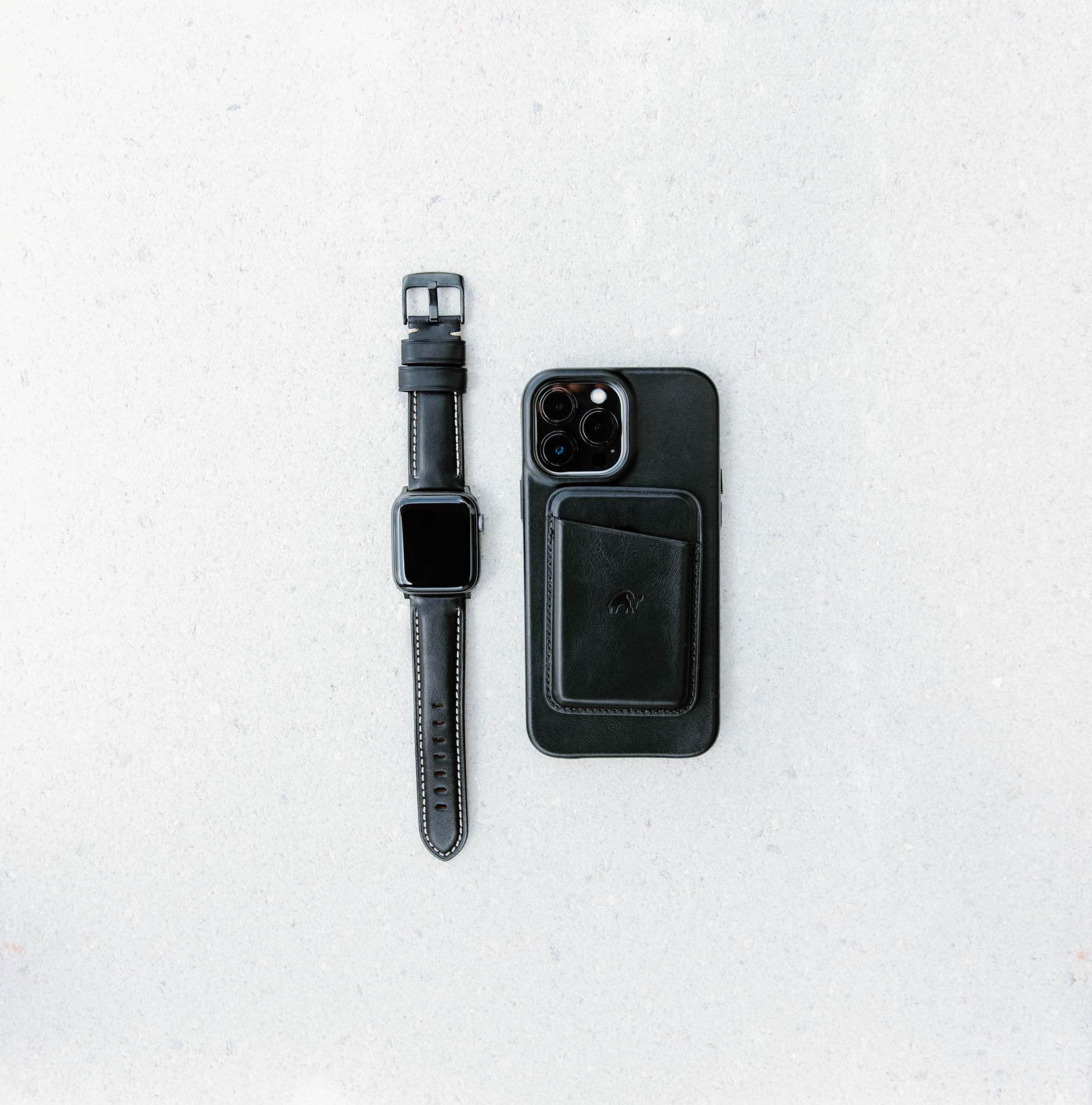 Black leather Apple Watch strap next to a black phone case on a gray surface.