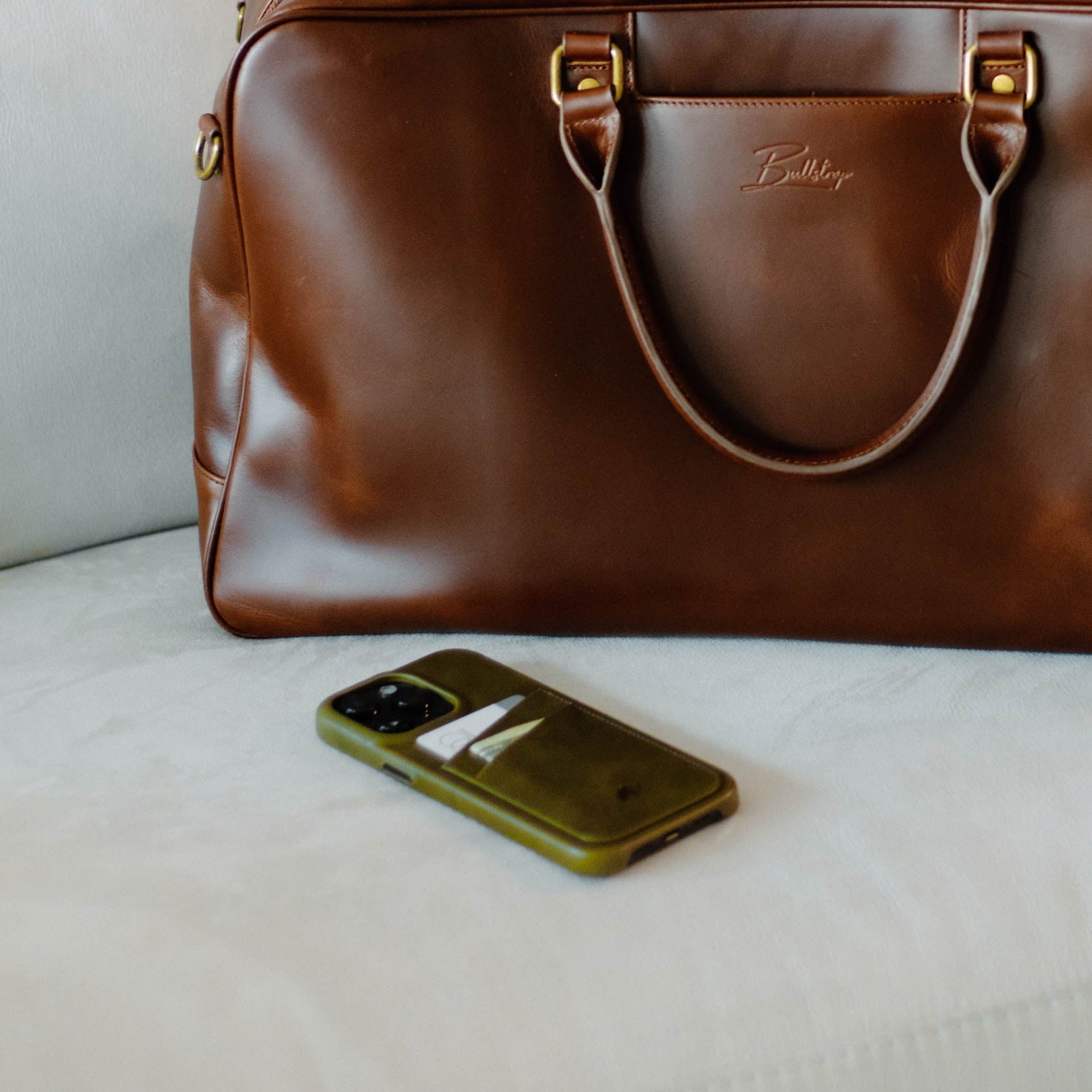 Brown leather duffel-style portfolio bag embossed Bullstrap, green phone with card in case on white surface