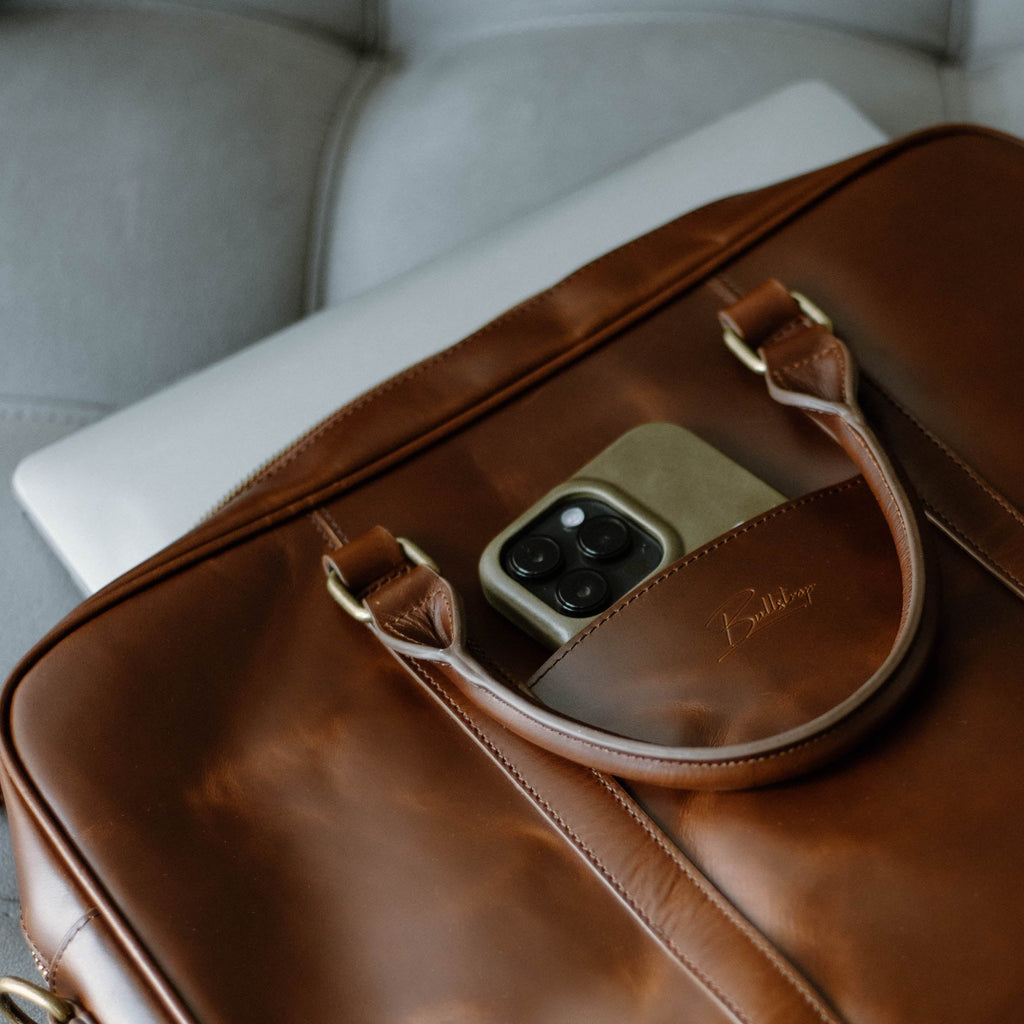 Brown leather briefcase with top handles, phone camera peeking from front pocket and a laptop partially inside
