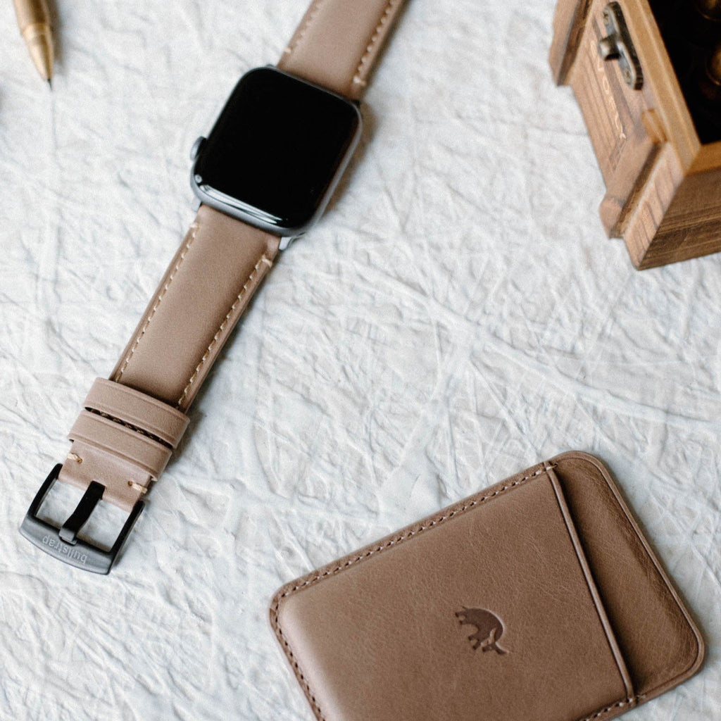 Leather Apple Watch Strap in Dune color, displayed with a wallet and pen on a textured surface.