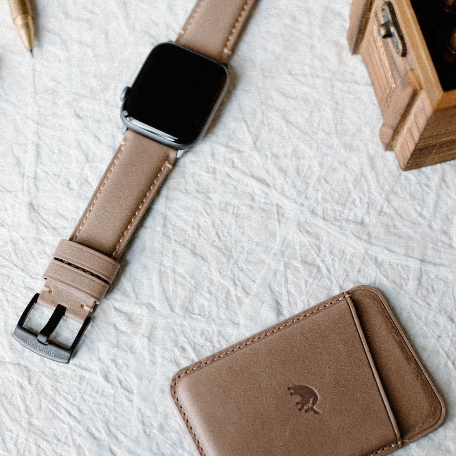 Leather Apple Watch Strap in Dune color, displayed with a wallet and pen on a textured surface.