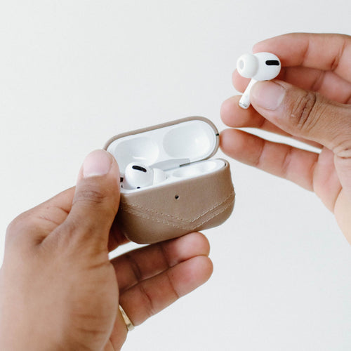 Two hands holding a tan leather AirPods charging case while removing a white wireless earbud.