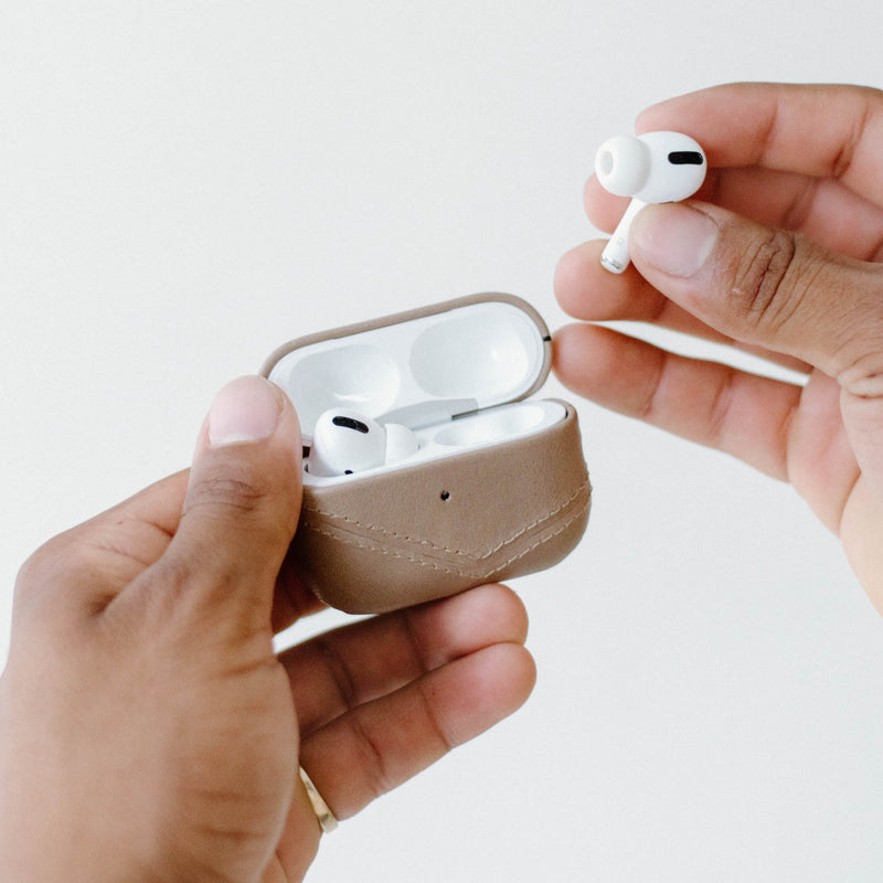 Two hands holding a leather AirPods charging case in DUNE while removing a white wireless earbud.