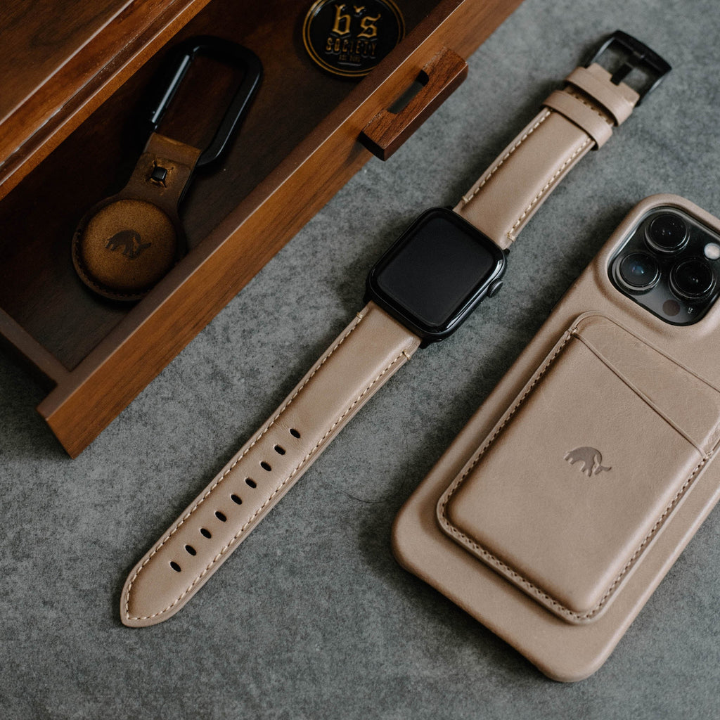 Leather Apple Watch Strap in Dune color, shown with a matching phone case and keychain on a gray surface.