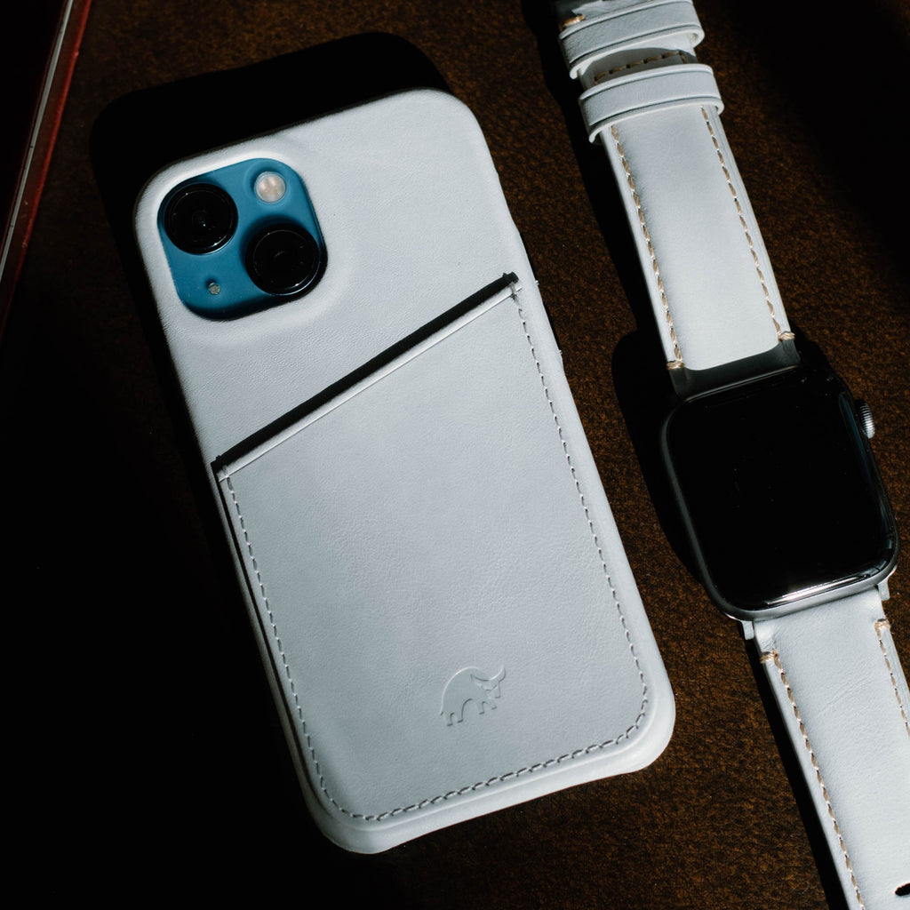 White leather iPhone case with rear card pocket and embossed elephant logo beside matching white leather smartwatch.