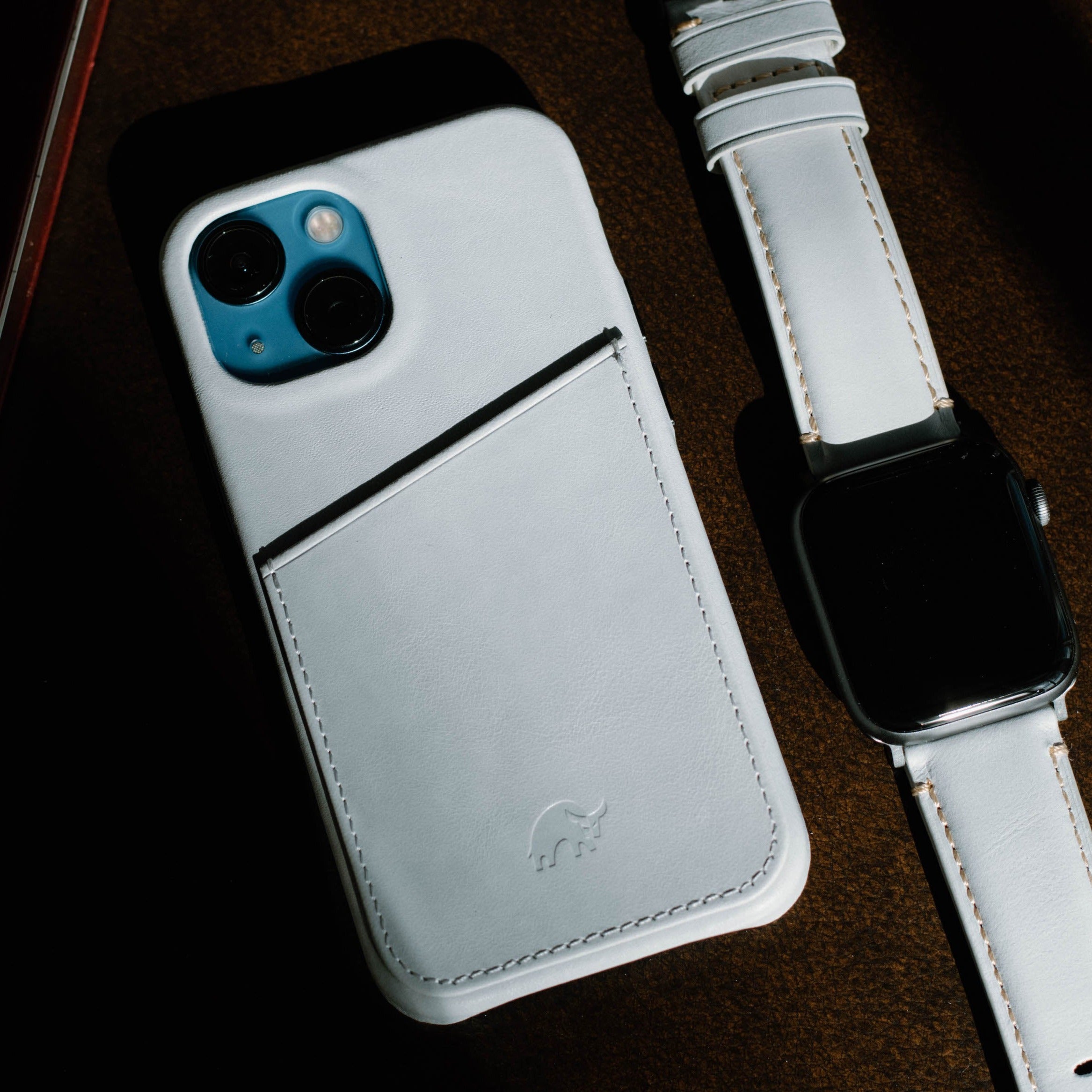 White leather iPhone case with rear card pocket and embossed elephant logo beside matching white leather smartwatch.
