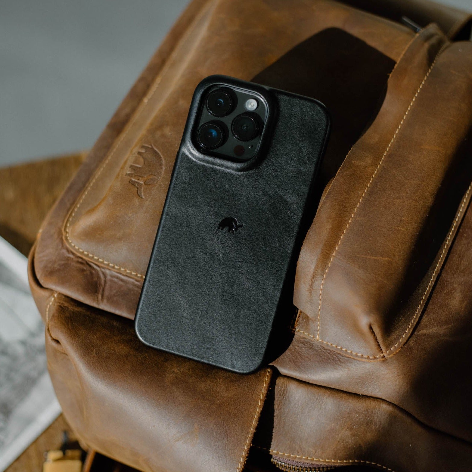 Black leather iPhone case with embossed logo resting on a brown leather backpack