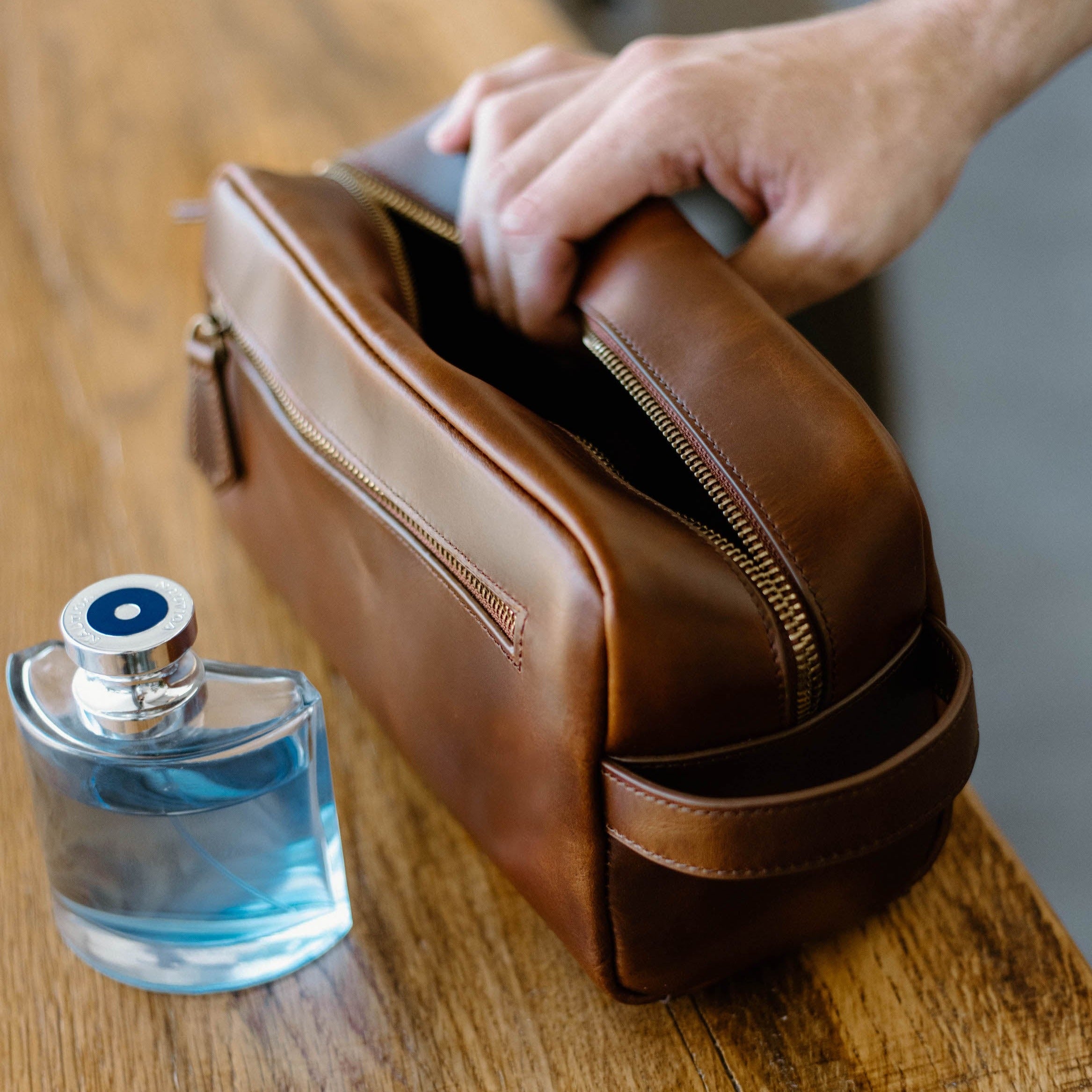 A hand reaching into a brown leather toiletry bag next to a blue bottle.