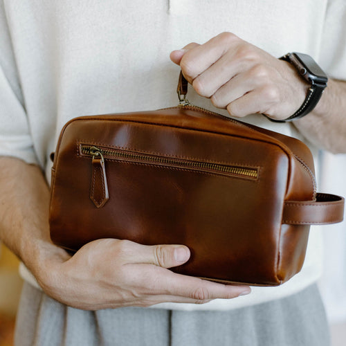 A person holding a brown leather toiletry bag with a zipper and handle.