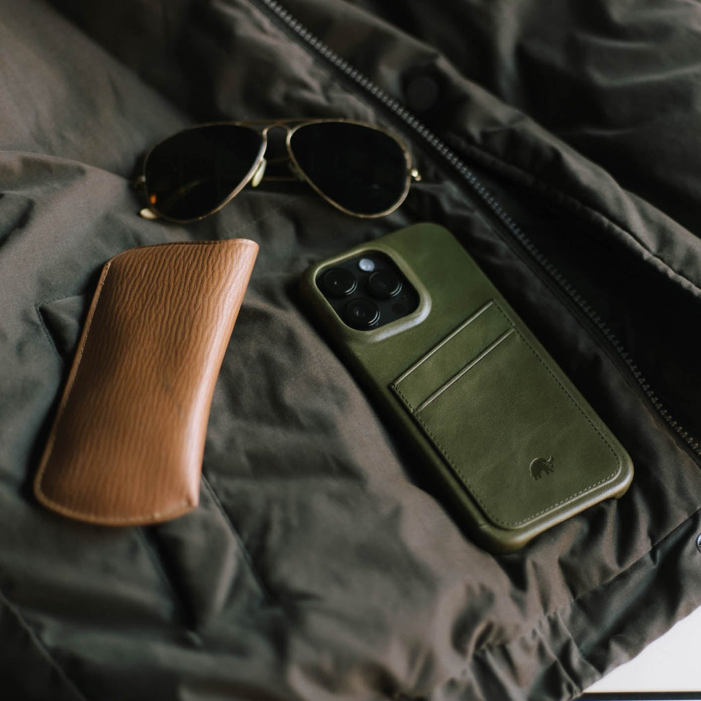 Green leather phone wallet case with two card slots on a dark jacket, beside a brown glasses case and aviator sunglasses