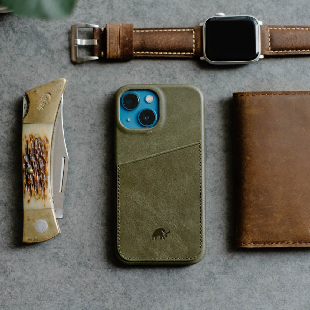 Olive-green leather phone wallet with diagonal card pocket and mammoth logo, shown with watch, knife, and brown notebook
