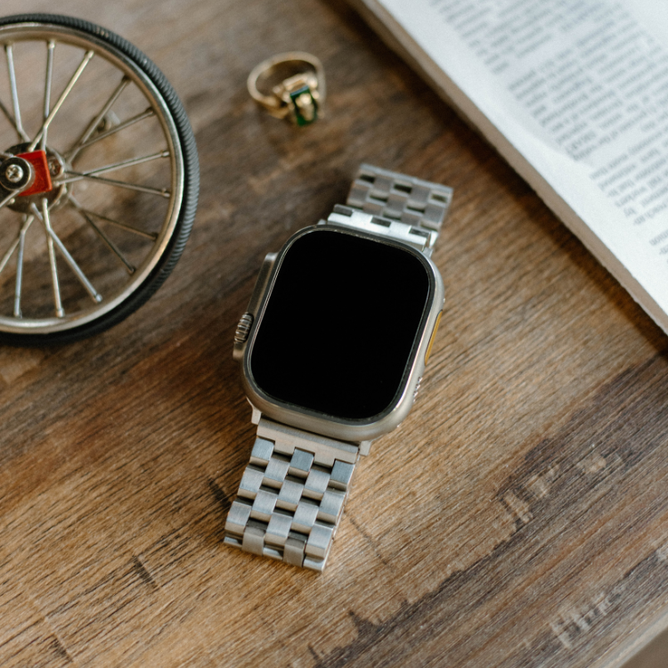 Stainless steel smartwatch with metal link band on a wooden table beside a miniature wheel, ring, and folded newspaper.