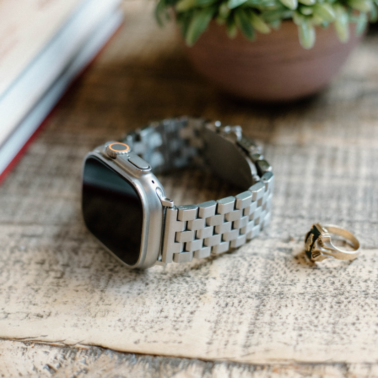 Silver smartwatch with metal link band and dark screen on a weathered wood table, gold ring beside it