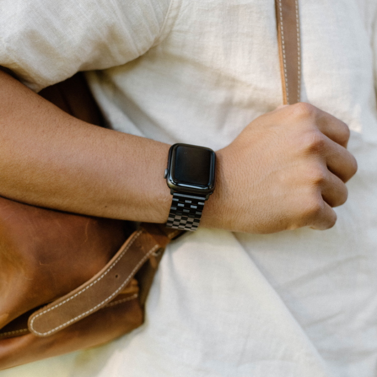 A person wearing a black metal Apple Watch strap on their wrist, with a brown bag in the background.