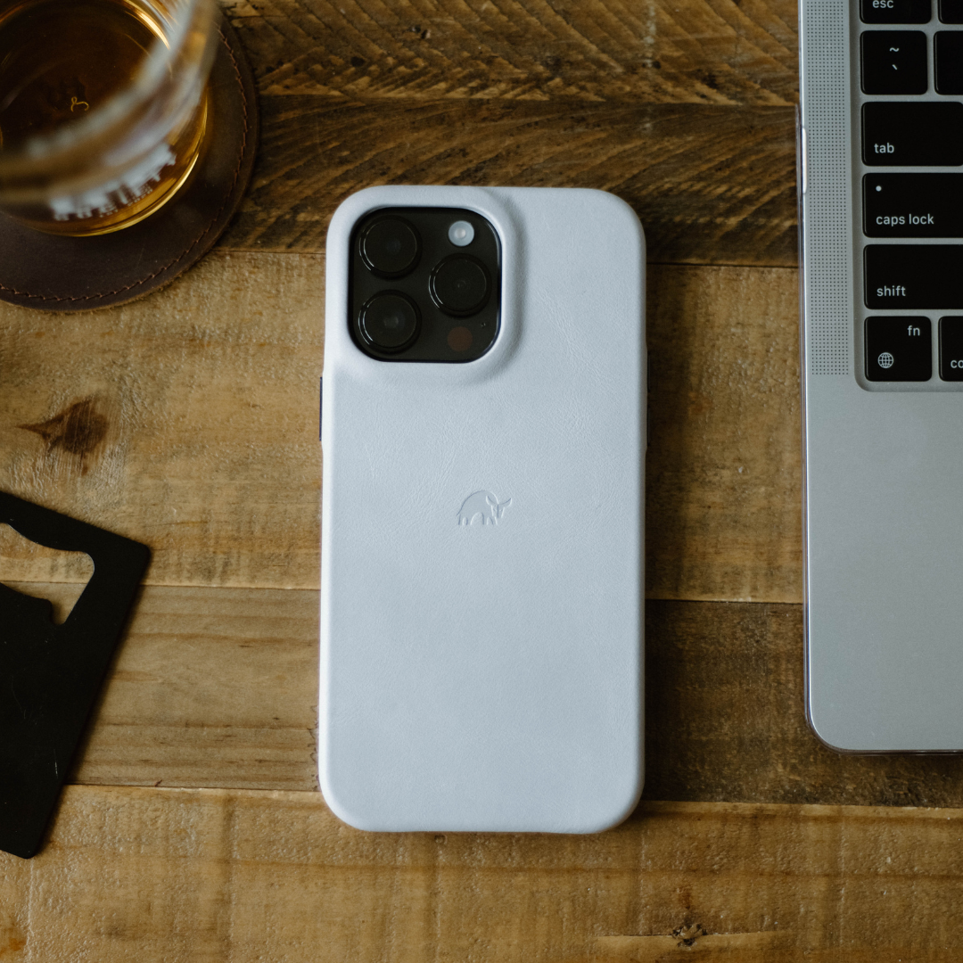 A white minimalist iPhone case on a wooden table next to a laptop and a drink.