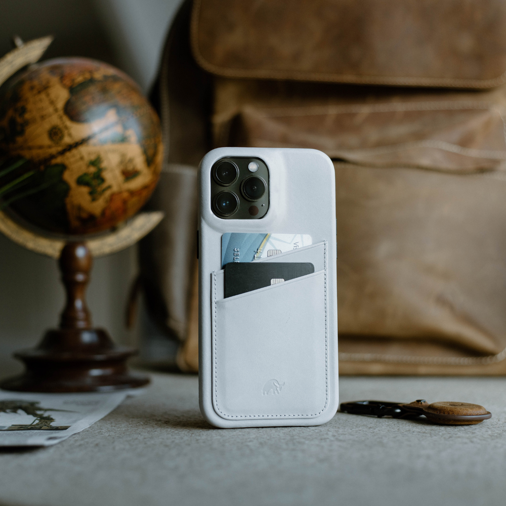 Minimalist white iPhone case with cards in pocket, on a table with a globe and backpack in background